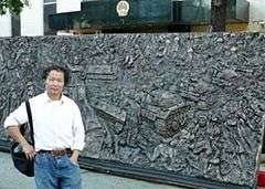 Asian male wearing white shirt and jeans in front of a bronze relief sculpture depicting people, tanks and a statue holding a torch; building with an official Chinese Government emblem is in the background