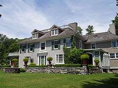 A large gray stucco house with white trim and green shutters. A low stone wall frames garden plantings around a patio in front of the house.