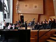 At right a woman in black stands in front of many children dressed in black, sitting in three rows of chairs on a large stage. In the background musicians play a grand piano, drums and guitars. At left a woman stands at a microphone, with several adults seated nearby. The stage opens to the outside at far left.