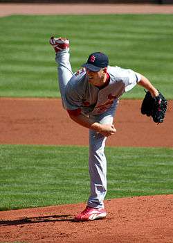 A man in a gray baseball uniform with red trim and navy baseball cap follows through after throwing a baseball from a dirt mound on a grass field with his right hand. His cap has an interlocked "StL" on the front in red type trimmed with white, and he has a black baseball glove on his left hand.