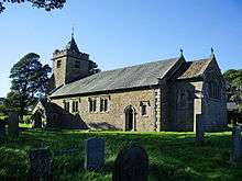 A small church seen from the southeast. The tower has a small pyramidal spire, a porch protrudes from the south wall, and the chancel to the right is short and slightly lower than the nave.  Gravestones in shadow are in the foreground