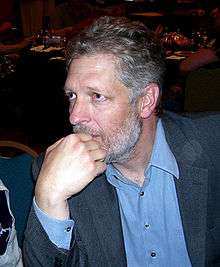 A Caucasian male with white hair/stubble rests his fist under his chin. He is wearing a blue suit. Part of another man also wearing a blue suit is sitting next to him.