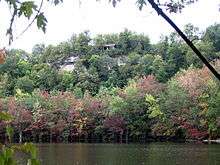 A cliff with a small shelter at its top, viewed from across a lake. The leaves in the trees growing from the slopes are green, pink, purple, and red.