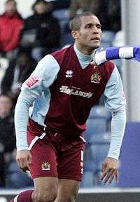 A man with very short hair who is wearing a blue top, white shorts and claret and blue striped socks. He is standing on a grass field.