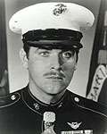 Head of a white man with a thin mustache wearing a white peaked cap with black visor and a dark military jacket with a medal hanging from a ribbon at the neck.