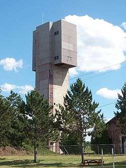 A large grey building, wider at the top that towers over the pine trees in the foreground