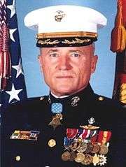 Head and shoulders of a white man wearing a white peaked cap and a dark military jacket with ribbon bars on the right breast, two rows of medals hanging from ribbons on the left breast, and a star-shaped medal hanging from a light-blue ribbon at the neck. Behind him are an American flag and a red and yellow flag.