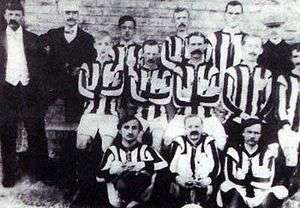 11 men in 3 rows, dressed in black-and-white striped football kit with 3 other men in suits