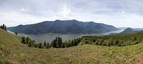 A wide river curves gently at the base of a mountain range. A meadow in the foreground gives way to an evergreen forest and then to the river. In the background, a layer of thin clouds veils a blue sky.