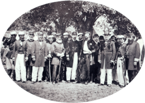 A photograph showing a large group men, most of whom wear uniforms, standing beneath shade trees