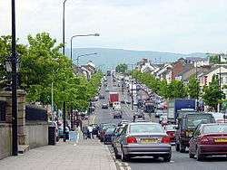 Four-lane street busy with vehicle traffic, with a central island, wide sidewalks, with two-story houses. In the distance, the 1700 ft mountain, Slieve Gallion.