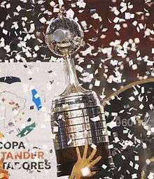 A silver trophy with a wooden base being lifted by a hand, amidst flying white paper pieces