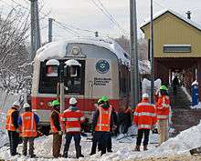 Eight men wearing neon orange jackets or vests with reflective stripes and green or white hard hats, most with their backs to the camera, stand in a ragged row in snow across the bottom of the image. In front of them is the front of a gray rail passenger train with a wide red stripe around its lower section between two gray square metal columns that rise to the top of the image. The words "Metro-North Commuter Railroad" and the Connecticut state seal are visible on one side of the front.  Its roof is covered with snow. In the rear right is a dull yellow off-center snow-covered gabled canopy with the words "New Canaan" in the field sheltering a concrete platform.