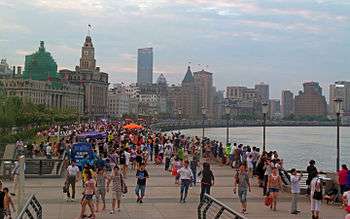 A wide curving walkway along a body of water to the right, crowded with many people and some large orange umbrellas underneath a sky colored by sunset. To the left of the walkway are light brownish-grey stone buildings in various late-19th and early-20th-century architectural styles, with taller buildings in the distance.