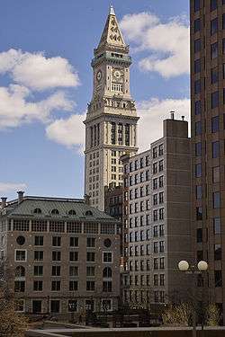 Distant, ground-level view of white limestone tower. The building is topped with a pyramidal-cap, and a large, 4-faced clock is visible just below the main roofline