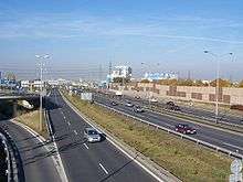 A view of the motorway from a flyover, showing two carriageways and access ramp to the left, with a noise barrier in the background.