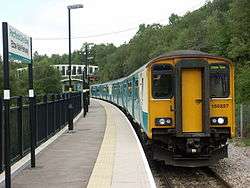 Four carriage train, on single curved track, at station. Steep wooded bank on one side of the track. Platform to the other. Station sign, in Welsh above English, in the foreground. Modern bridge over the track in the background.