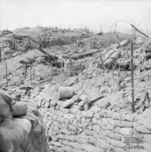 A black and white photo of damaged field defences in the daylight. A deep sandbag-lined trench runs from the bottom right to the bottom left away from the camera, while further up the slope to the right disturbed earth, damaged star pickets and wire entanglements and other debris is evident below the ridgeline.