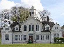  Ornate, symmetrical, wooden house with two smaller wings with decorative engaged columns. The pitched roof has gables. The windows are paned and the front door has columns on either side.