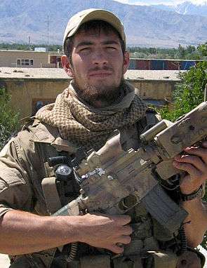 A bearded young man wearing a hat on the top of his head as well as a scarf around his neck while squinting. He wears a vest on his torso and is holding a carbine in his hands, the barrel of it facing upward. Behind him are beige-colored dwellings, shipping containers, and the peaks of snow-capped mountains.