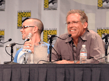 Two men seated in front of a table with microphones. The San Diego Comic Con logo can be seen in a panel behind them.