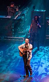 A man at the front of a stage holding a guitar and standing behind a microphone stand. Behind him are two men, one wearing sunglasses and holding a violin and the other in a striped shirt playing keyboards. The stage is lit from behind by a blue light that casts the shadows of leaves and thin branches.