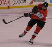 An ice hockey player is balanced on one foot with his stick in the air having just taken a shot. He is wearing an orange uniform with white bands on each arm and '54' on the sleeve. He is also wearing a black helmet.