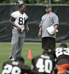 Candid photograph of Shamash wearing a long-sleeved grey t-shirt bearing an NFL logo and a brown baseball cap bearing a Cleveland Browns logo while standing on a football field talking with a football player