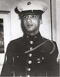 Head of a young black man looking off to the side, wearing a white peaked cap with black visor and dark military jacket.