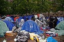 A group of small tents in a park with green trees, concrete sidewalks and falling snow, some of which has begun to accumulate on the tents