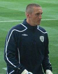 A man with very short hair is wearing a dark jacket. He is standing on a grass field.