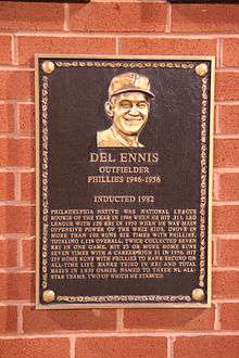 A dark bronze plaque with a man's face at the top and gold writing beneath, mounted on a brick wall