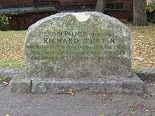 A stone gravestone with a curved top, with several lines of inscription.  Trees, grass, and a wall are visible in the distance.