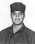 Head and shoulders of a smiling young white man wearing a cloth army cap and a plain buttoned-down shirt of the same color.