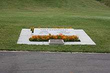 A grave marked by an angled slab of marble engraved with the names of Diefenbaker and his wife and surrounded by plantings of small marigolds, and an additional plaque in bronze. Beyond it a lawn stretches away.