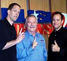 Three men all face forward and are smiling. Two of the men are extending their pointer fingers while the third man is giving a thumbs up gesture.