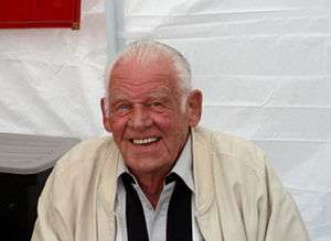 A manwearing a vanilla-colored jacket, with a white collared dress shirt and a black scarf around his neck