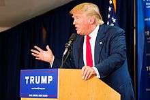 Trump speaking behind a brown wooden podium, wearing a dark blue suit and a red tie. The podium sports a blue "TRUMP" sign.