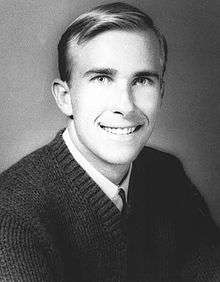 Head and shoulders of a young white man with a bright smile and neatly combed hair, wearing a dark sweater over a shirt and tie.