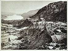 Lake beside snowy mountains with railroad construction sheds in foreground.