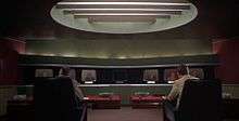 A room fitted out in futuristic décor contains a panel of monitor screens, each presenting the face of a teleconference delegate. In the foreground of the shot, men address the delegates on the monitors.