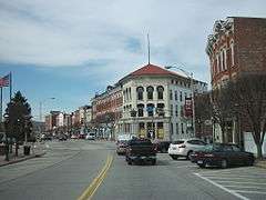 A row of two-story commercial brick buildings, some painted, on a street curving away from the viewer to the right and left. In front of them is a road, some short bare trees, and diagonally parked cars.