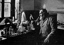 A bearded old man holding up a test tube. He is sitting at a table by a window. The table is covered with many small bottles and test tubes.