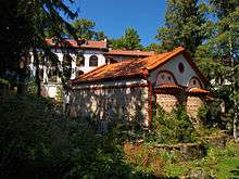 Apse view of a late medieval stone church with a tiled roof and a residential building in the background, all located in a forested area