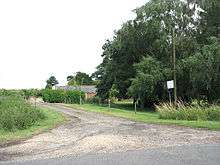 Small farm track leading off a mintor road.  behyind a low hedge some single story brick farm buildings peep.  A huge tree ominates the right side of the picture