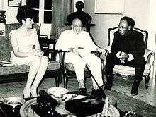 An elderly, smiling Du Bois sits in a chair, flanked by a man and woman also seated and smiling.