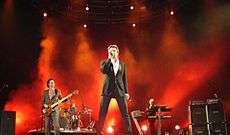 Four men performing on a stage in front of a red mist. In the foreground a white man sings into a microphone. Another male can be seen playing the guitar