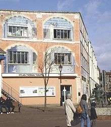 A wide three storied building with windows to the upper two stories and an entrance on the ground floor. A statue of Thomas sits outside.