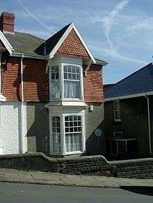 On a hill street stands a two storied semi-detached house with bay windows to the front and a sloped tiled roof with a chimney.