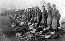 A line of soldiers standing behind tents with their army gear neatly laid out ready to be inspected. The line of soldiers continues into the distance were buildings can be seen the background.